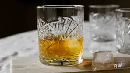 Kryształowa szklanka w stylu vintage wypełniona whisky, ustawiona na drewnianej desce z kostką lodu obok.